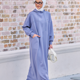 Woman wearing a light purple outfit with a headscarf against a brick wall.